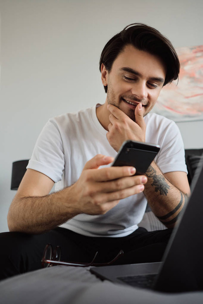 Beyaz t-shirt genç gülümseyen esmer adam mutlu modern evde dizüstü bilgisayar ile yatakta oturan cep telefonu kullanarak - Fotoğraf, Görsel