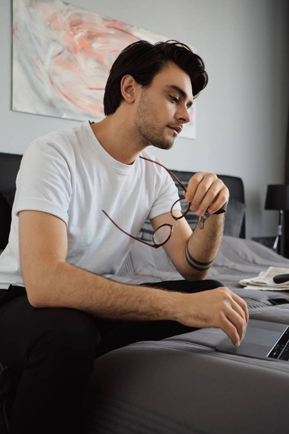 Молодий привабливий втомлений брюнетка чоловік у білій футболці сидить на ліжку з окулярами в руці, вдумливо працюючи на ноутбуці в сучасному будинку
 - Фото, зображення