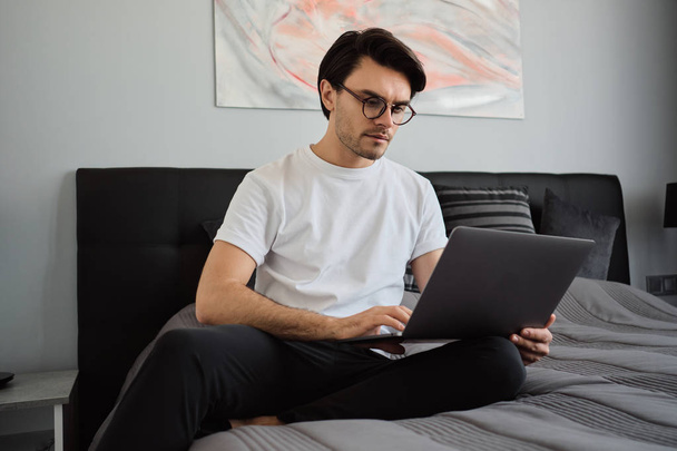 Jovem atraente morena séria homem em t-shirt e óculos sentado na cama em casa moderna cuidadosamente trabalhando no laptop
  - Foto, Imagem