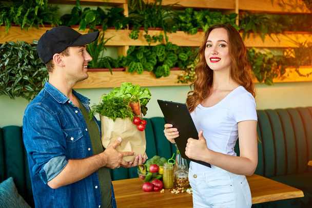Courier service worker delivering fresh food to a happy woman client signing some documents on the kitchen at home. Online grocery shopping from the internet - shop - Фото, изображение