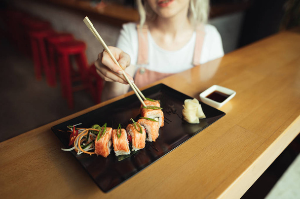 Background. Woman eats appetizing sushi roll in Japanese restaurant, takes rolls with chopsticks, cropped photo from above, Girl hands take sushi rolls with chopsticks from sushi plate. - Foto, afbeelding