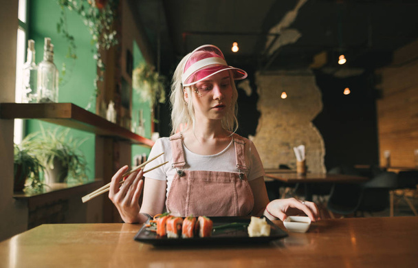 Menina atraente senta-se em um acolhedor restaurante japonês com um prato de rolos de sushi e molho de soja, segura pauzinhos, olha para baixo, usa um boné rosa e sundress. Restaurante asiático conceito
. - Foto, Imagem