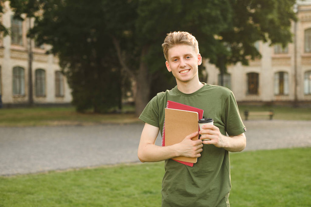 Νέος χαμογελαστός άντρας μαθητής στέκεται στο γκαζόν στο κολλέγιο πάρκο κρατώντας μακριά καφέ και σημειωματάρια. Επιτυχής χαρούμενο Πανεπιστήμιο φοιτητής με ένα πλατύ χαμόγελο κοντά στην πανεπιστημιούπολη με καφέ και βιβλία. - Φωτογραφία, εικόνα