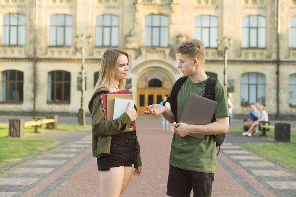 若い魅力的な学生のカップルは、ノートパソコン、書籍、ノートを持って大学の近くの公園で屋外で話しています。キャンパスの近くで会話をしている2人の大学生. - 写真・画像