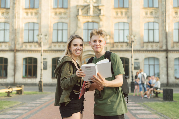 Szczęśliwy wesoły dwóch studentów na kampusie uniwersyteckim trzymając książkę patrząc na kamerę. Atrakcyjne pozytywne studentów para stoi przed budynkiem kolegium z podręcznym. - Zdjęcie, obraz