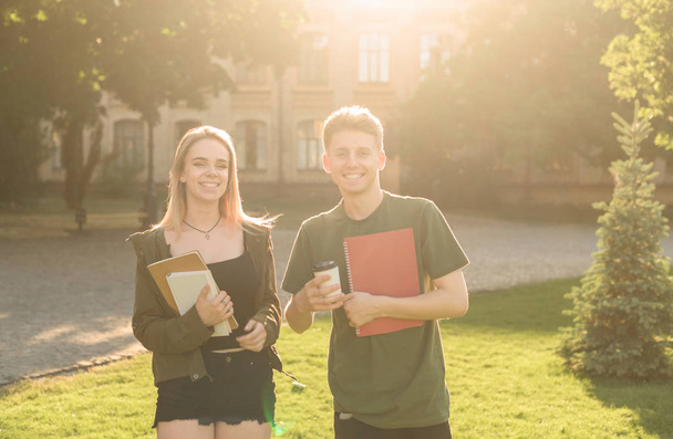 Twee opgewonden studenten met goedgekeurde examens in de buurt van de Universiteit houden de notebooks en neem weg koffie kopje. Paar studenten in het Park in de buurt van het College glimlachend en gevoel onzorgvuldig. - Foto, afbeelding