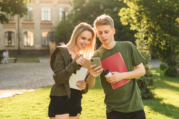 Deux étudiants heureux vérifiant le contenu du téléphone intelligent debout dans le parc de l'université tenant des livres et des cahiers. Quelques étudiants joyeux sur le terrain de l'université regardant le smartphone
. - Photo, image