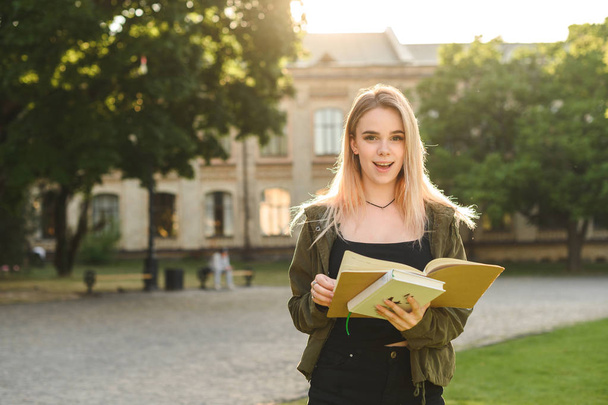 Έκπληκτος νεαρή όμορφη γυναίκα μαθητής που ποζάρει στο κολλέγιο πάρκο σε εξωτερικούς χώρους κρατώντας βιβλία και σημειωματάρια. Ελκυστικός χαριτωμένος μαθητής ευχάριστα έκπληκτος από τους θετικούς βαθμούς στο Πανεπιστήμιο. - Φωτογραφία, εικόνα