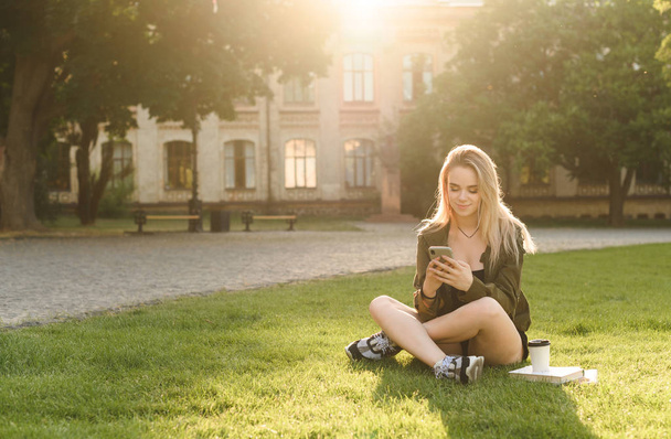 Young college student girl using smartphone while sitting on the grass in the college garden. Positive female student near the university building sitting in the park texting on her phone. - Foto, Imagem