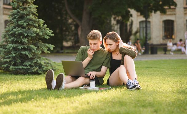 Attentive teen students couple searching content, commenting on laptop sitting on grass in university park. Concentrated students looking at laptop screen, examining information in college garden. - Φωτογραφία, εικόνα