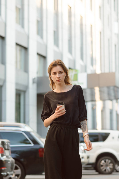 Portret poważnej eleganckiej kobiety patrząc na aparat ubrany w szarą bluzkę i spodnie trzymając kawę przed wieżowcem. Młoda stylowa dziewczyna spaceru w pobliżu parkingu w miejskiej scenerii z filiżanką kawy. - Zdjęcie, obraz