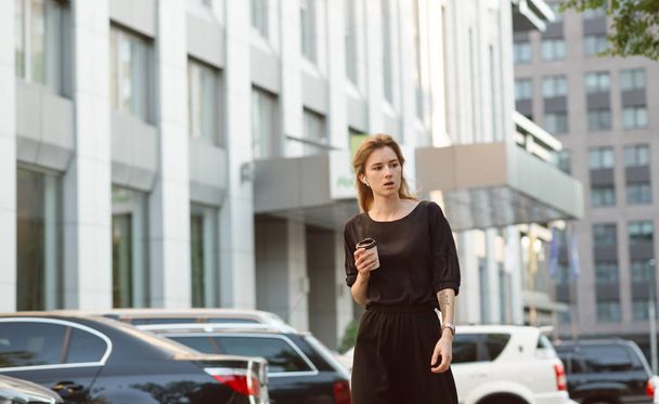 Portrait d'une jeune femme d'affaires sérieuse marchant près des bâtiments urbains, tenant une tasse de café et regardant de côté. Femme d'affaires professionnelle buvant du café, en déplacement style de vie extérieur
. - Photo, image