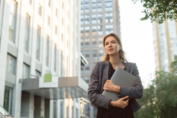 Portret całkiem bizneswoman w Smart casual ubrania na zewnątrz budynku biurowego. Młoda kobieta pracownik biurowy trzyma laptopa, uśmiechając się w jej bezprzewodowych słuchawek w pobliżu centrum biznesowego. - Zdjęcie, obraz