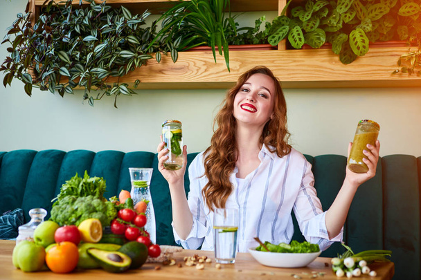 Νεαρή γυναίκα που πίνει χυμό και φρέσκο νερό στο όμορφο εσωτερικό με πράσινα λουλούδια στο φόντο και φρέσκα φρούτα και λαχανικά. Έννοια της υγιεινής διατροφής. Χορτοφαγικό γεύμα και μενού αποτοξίνωσης - Φωτογραφία, εικόνα