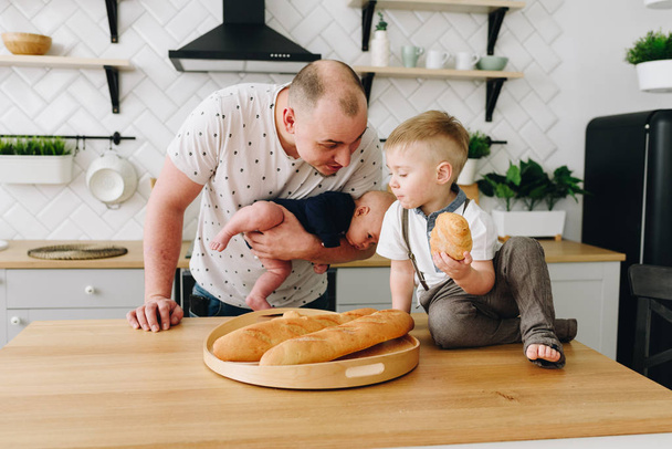 Jovem pai caucasiano conversando com seu filho e segurando um bebê na cozinha
 - Foto, Imagem