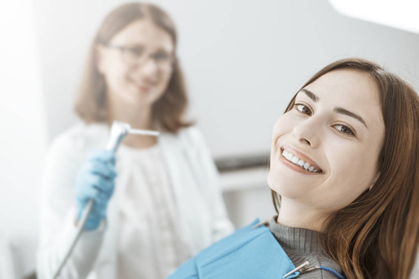 Genç bir kadın bir diş sandalyesinde oturur ve beyaz ve sağlıklı dişler ile gülümsüyor. Doktor ve mutlu hasta.  - Fotoğraf, Görsel