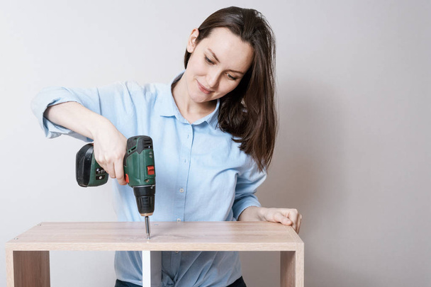 Confident European woman collects wooden furniture with electric screwdriver in hand - Photo, Image