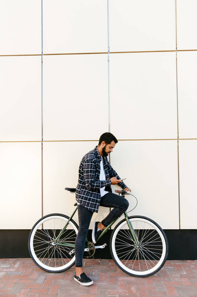 Side view of stylish man sitting on bike with cell phone, outdoors - Valokuva, kuva