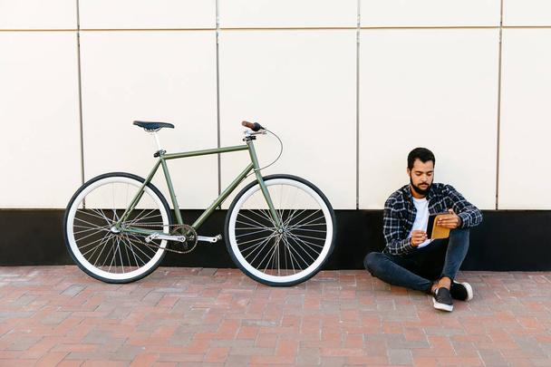 Bearded man sitting with tablet near the bike, outdoors. - Photo, Image