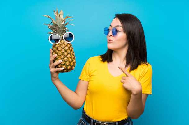 Joven mexicana sobre fondo azul aislado sosteniendo una piña con gafas de sol
 - Foto, imagen