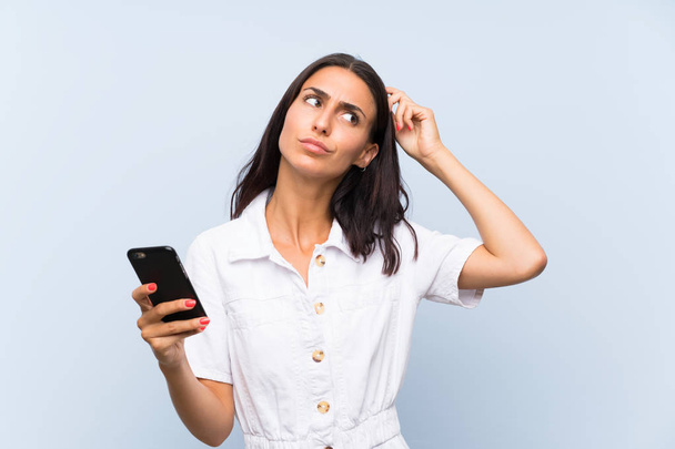 Молода жінка з мобільним телефоном над ізольованою синьою стіною має сумніви і з плутаниною вираз обличчя
 - Фото, зображення