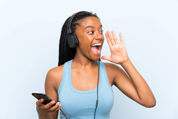 African American teenager girl with long braided hair listening music with a mobile shouting with mouth wide open - Zdjęcie, obraz