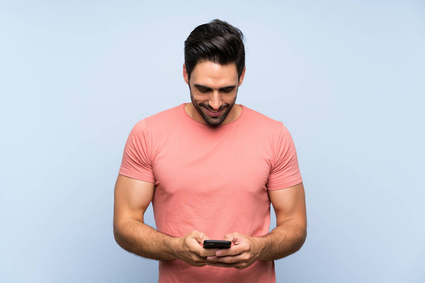 Pohledný mladý muž v růžové košili přes izolované modré pozadí, které posílá zprávu s mobilním - Fotografie, Obrázek