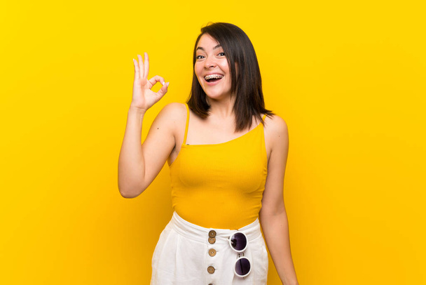 Young Mexican woman over isolated yellow background surprised and showing ok sign - 写真・画像