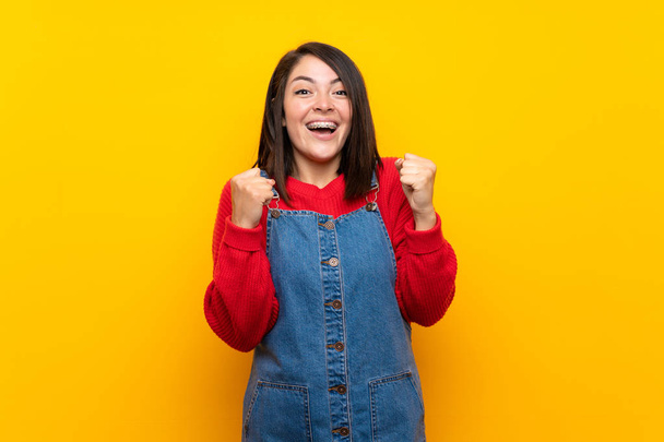 Young Mexican woman with overalls over yellow wall celebrating a victory in winner position - Photo, Image