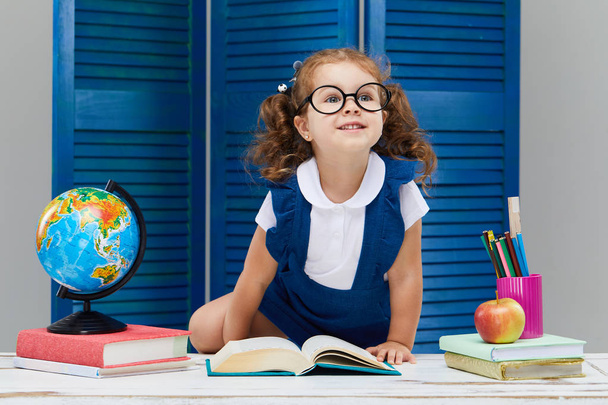 Smart toddler kid. Back to school and happy time! Cute industrious child in glasses is sitting at a desk indoors. Kid is learning in class on background of blue wall. Girl reading the book. - Photo, image