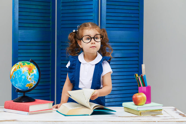 Smart toddler kid. Back to school and happy time! Cute industrious child in glasses is sitting at a desk indoors. Kid is learning in class on background of blue wall. Girl reading the book. - Photo, image
