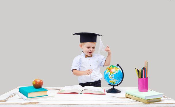 Clever and smart child, prodigy. A little boy in a hat is studying geography on a globe. Examines a map through a magnifying glass. Sitting at a desk with books on a gray isolated background - Photo, Image