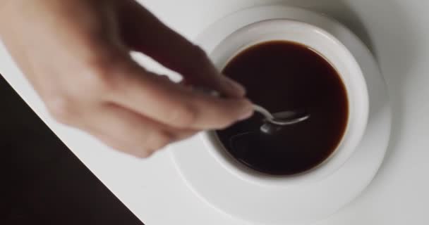 Вид зверху руки перемішуючи цукор в чашці гарячої кави з ложкою на білому столі. Пити і насолоджуватися смачною кавою американо з білого кубку. Красивий крупним планом вид з ранкової кави в кафе
 - Кадри, відео