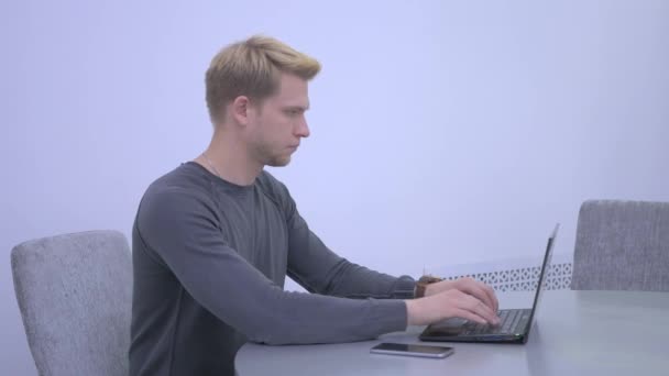 Busy man in grey shirt sitting on the chair on the table and typing on his laptop. Serious man looking in the screen of computer and moving his fingers on the board very quickly. The man looking in camera and smiling. - Кадры, видео