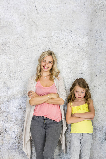 pretty daughter with blonde young mother standing with crossed arms on grey wall background  - Фото, зображення