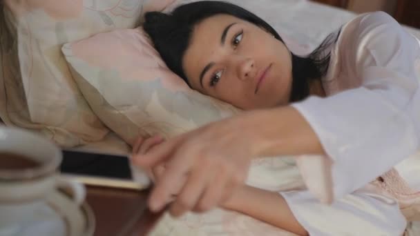 Close-up. The camera focuses on a young pretty woman, who's relaxing in the bed. The woman take a smartphone from a table near the bed and using it. The woman looking in the screen and pushes some keys on a screen. - Video