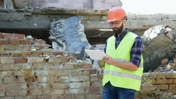 Um engenheiro construtor em um capacete está calculando o plano de demolição do edifício e olhando para o projeto de construção em um tablet
 - Filmagem, Vídeo