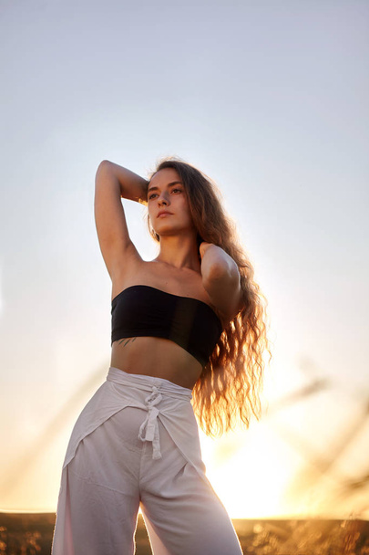 Portrait of beautiful young stylish girl in the sunlight at sunset. Posing.Fashion.Long haired,curly girl.She is cute.Beautiful woman is enjoying warm and sunlight. She is bathing in the sun - 写真・画像
