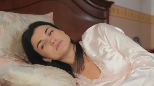 Young brunette woman laying in the bed. Beautiful brunette stand up and leaned on the arm in the bed. Pretty young woman takes a cup with tea in her hands from a wooden table and start drinking a tea. - Materiał filmowy, wideo