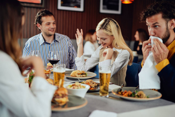Мультиэтнические друзья сидят в ресторане, пьют алкоголь, болтают и едят бургеры на ужин
. - Фото, изображение