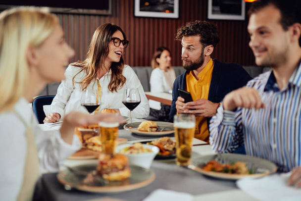 Amigos multiétnicos sentados en el restaurante, bebiendo alcohol, charlando y comiendo hamburguesas para cenar
. - Foto, imagen