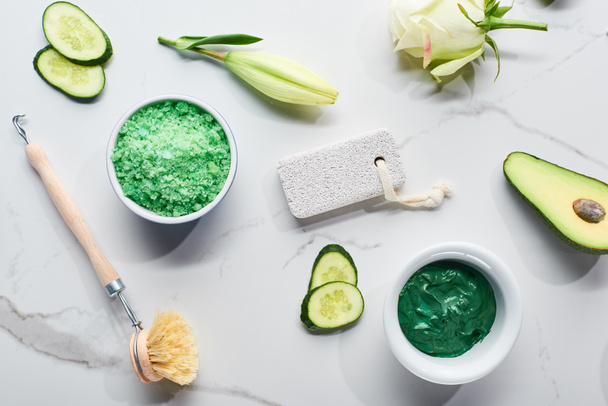 top view of body brush, pumice stone, bath salts and face mask near fresh avocado half, cucumber slices and flower on marble background - Photo, Image