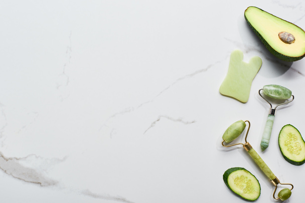 top view of facial spatula and rollers near avocado and cucumber on marble surface - Photo, Image
