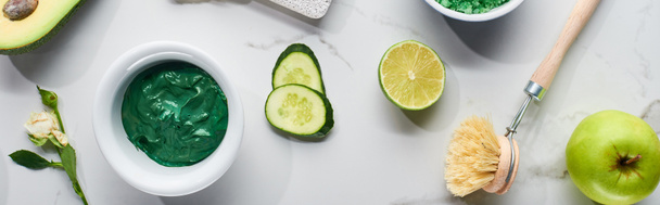 panoramische shot van natuurlijke schoonheid product en Body Brush in de buurt van komkommer schijfjes, limoen, avocado en groene appel op marmeren oppervlak - Foto, afbeelding