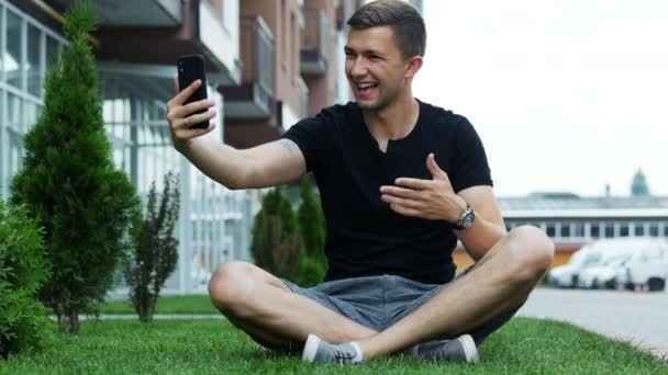 Pohledný mladý muž, který mluví v videokonverzaci přes mobil, sedí na trávě a mluví s přáteli - Záběry, video