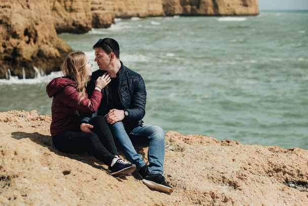 Щаслива молода пара обіймаючи і тримає руки на березі океану. Чоловік і жінка сидять разом на скелі. Усміхнена пара в чорних окулярів насолоджуючись відпочинком в Португалії - Фото, зображення