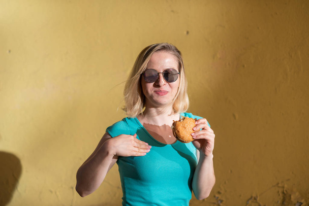 Retrato de una linda chica con un vestido verde caminando afuera y comiendo una hamburguesa en un cálido día de verano. Hermosa rubia en gafas de sol disfruta de comida chatarra en la pared amarilla en la calle. Hábitos alimenticios incorrectos
. - Foto, imagen