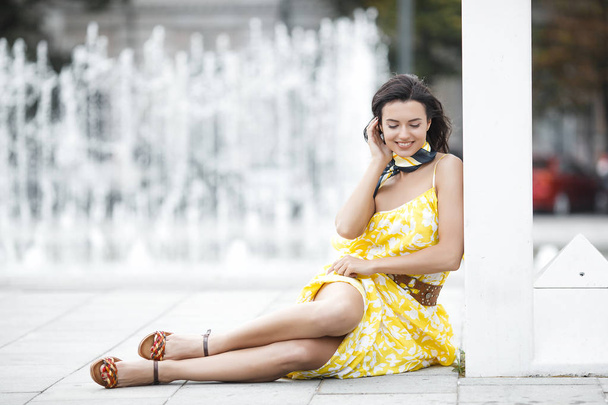 Vonzó fiatal nő a szökőkút közelében. Gyönyörű női szabadban. Hölgy sárga ruhában portré. - Fotó, kép