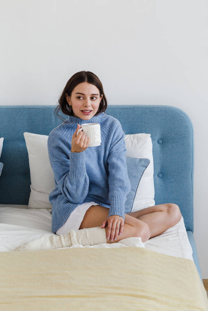  Girl in a blue sweater in hygge style interior with a cup of hot tea her hand sits on the bed - Foto, Imagen