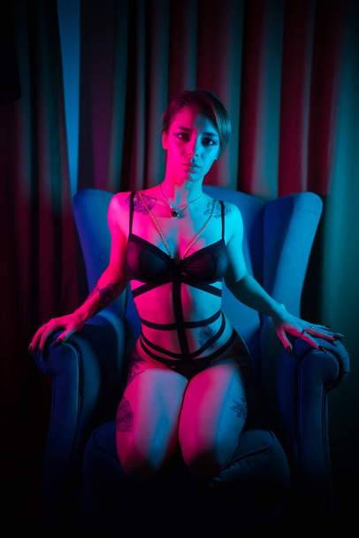 Молодая женщина с татуировками в сексуальном белье сидит в кресле в неоновом свете
 - Фото, изображение